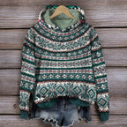 Pull-over à motifs nordiques verts et blancs suspendu sur un cintre en bois, accompagné d'un short en jean déchiré, sur fond de bois rustique.
