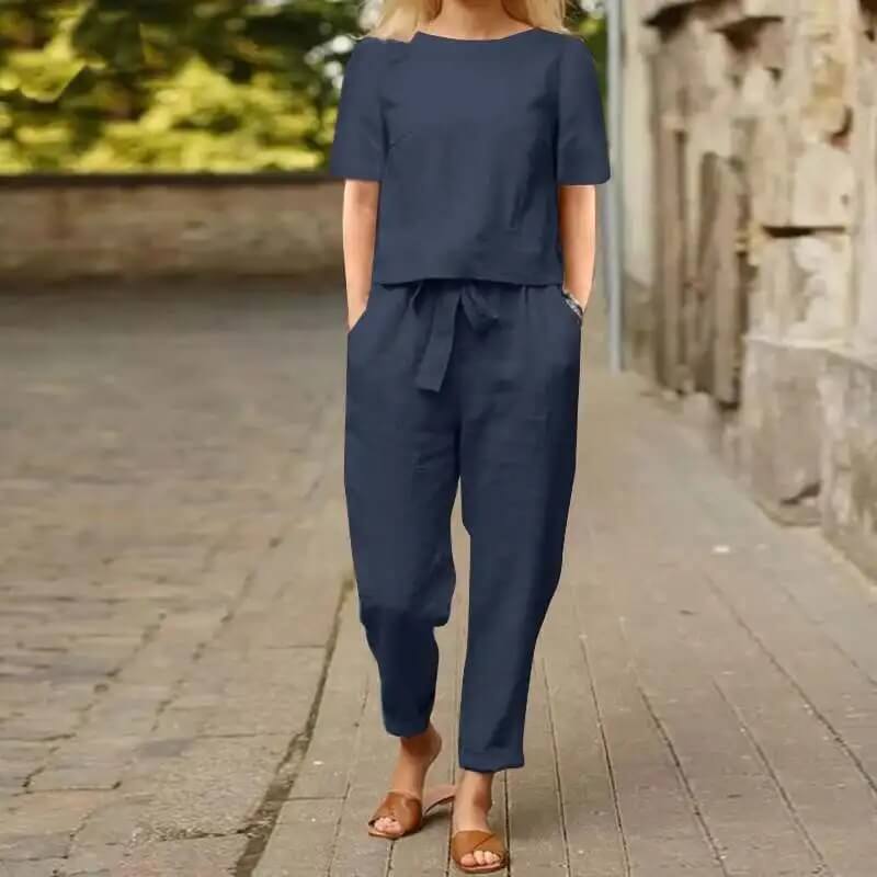 Femme en combinaison bleu marine, manches courtes, marchant sur un trottoir pavé, portant des sandales marron. Mode décontractée, tenue estivale élégante.