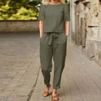 Femme en combinaison kaki, manches courtes, marchant sur un trottoir pavé, portant des sandales marron. Mode décontractée, tenue estivale élégante.