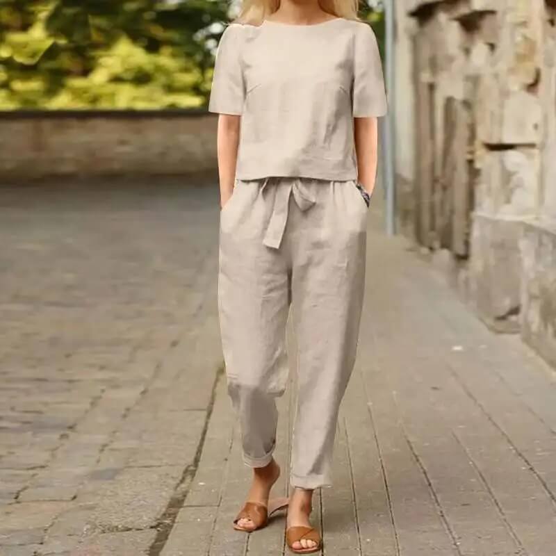 Femme en ensemble beige en lin, pantalon et haut à manches courtes, marchant sur une rue pavée. Mode décontractée, tenue estivale élégante.