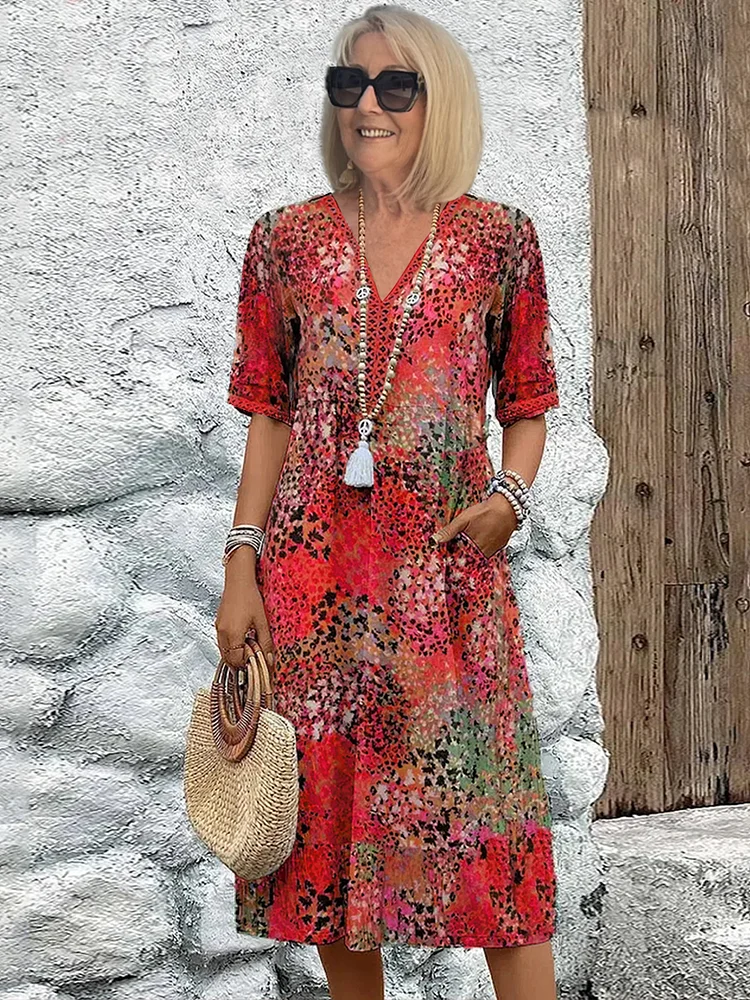 Femme souriante en robe d'été fleurie rouge, portant des lunettes de soleil et un sac en paille, posant devant un mur en pierre blanche.