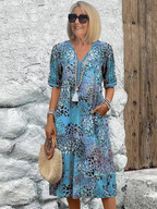 Femme souriante en robe bleue à motifs floraux, portant un sac en paille et des lunettes de soleil, posant devant un mur blanc et une porte en bois.