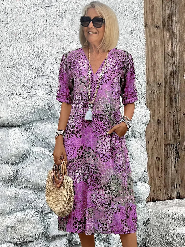 Femme souriante en robe violette à motifs floraux, portant un sac en paille et des lunettes de soleil, posant devant un mur texturé.