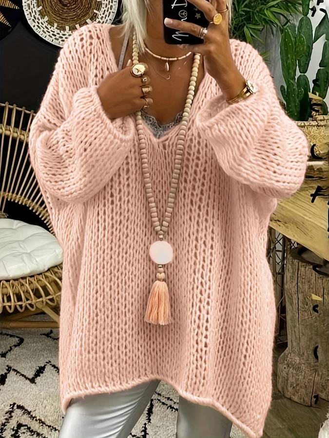 Femme portant un pull oversize rose en maille, collier long avec pompon, leggings argentés, décor intérieur bohème avec plantes et fauteuil en rotin.
