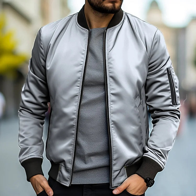 Homme portant un blouson bomber gris argenté, manches longues, col noir, sur un t-shirt gris, en extérieur urbain flou. Mode masculine moderne.