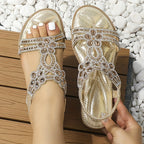 Sandales dorées ornées de strass sur pieds féminins, posées sur une terrasse en bois avec des galets blancs. Mode estivale élégante et chic.