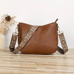 Sac à main en cuir marron avec bandoulière à motifs floraux, posé sur une table en bois clair, décoré de fleurs séchées en arrière-plan.