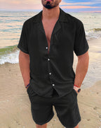 Homme en chemise noire sur la plage, tenue estivale élégante, mer en arrière-plan, mode masculine décontractée, coucher de soleil.