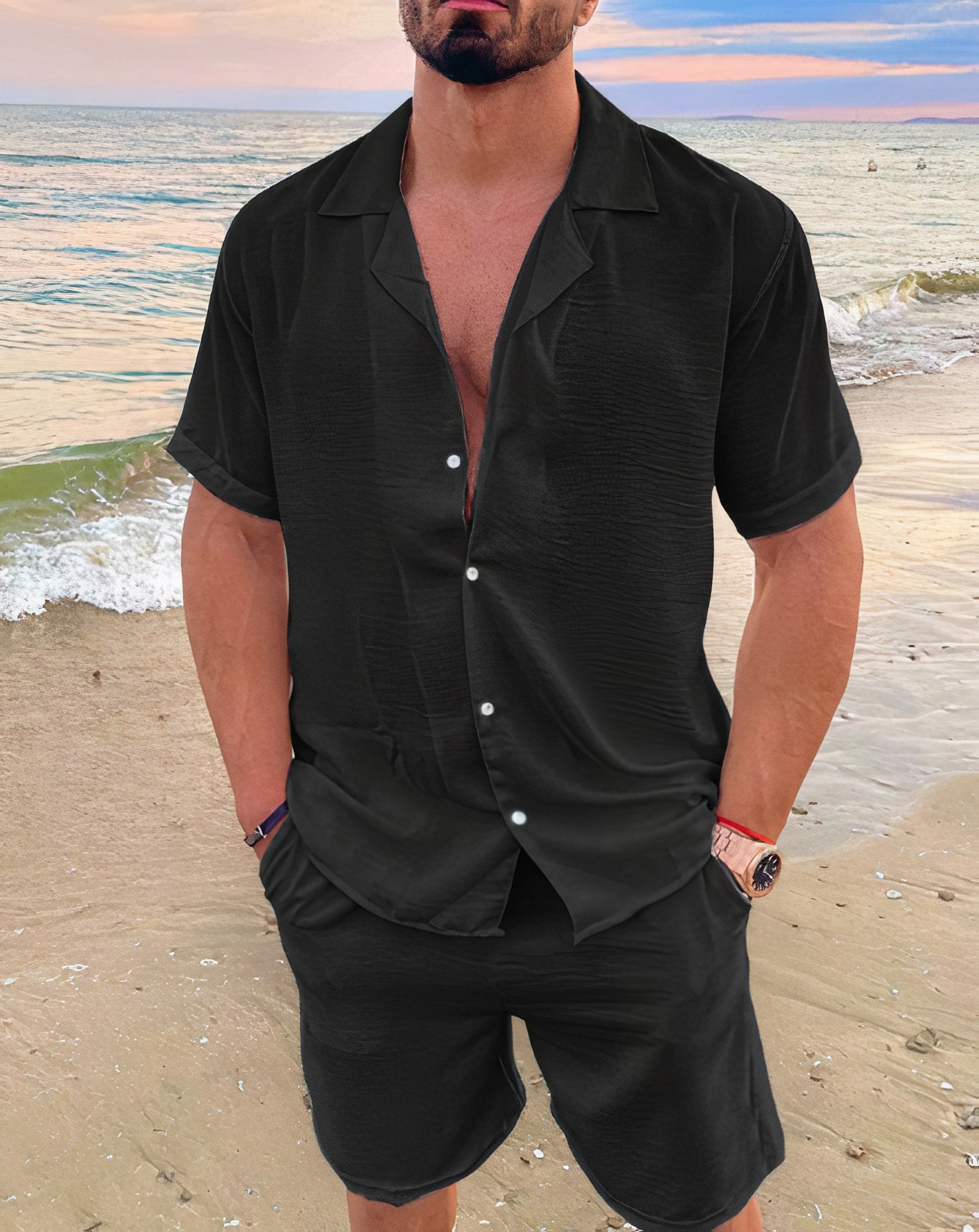 Homme en chemise noire sur la plage, tenue estivale élégante, mer en arrière-plan, mode masculine décontractée, coucher de soleil.