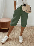 Femme portant un short vert kaki, t-shirt blanc, baskets blanches et sac à main en paille, posant sur un tapis beige. Mode décontractée chic.