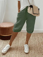 Femme portant un short vert kaki, t-shirt blanc, baskets blanches et sac à main en paille, posant sur un tapis beige. Mode décontractée chic.