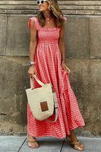 Femme en robe longue à carreaux rouges, portant un sac en paille et des lunettes de soleil blanches, posant devant un mur en pierre.