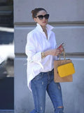 Femme élégante en chemise blanche, jeans décontracté, lunettes de soleil, tenant un sac à main jaune et un sac en papier, marchant en ville.
