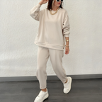 Femme en ensemble beige décontracté, sweat et pantalon assortis, portant des baskets blanches et des lunettes de soleil, posant contre un mur blanc.