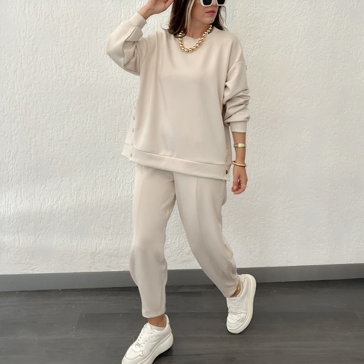 Femme en ensemble beige décontracté, sweat et pantalon assortis, portant des baskets blanches et des lunettes de soleil, posant contre un mur blanc.