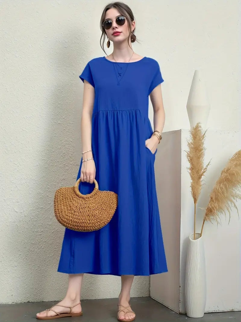 Femme en robe bleue élégante, portant un sac en osier, lunettes de soleil, sandales beiges, posant près d'un vase décoratif avec des pampas.