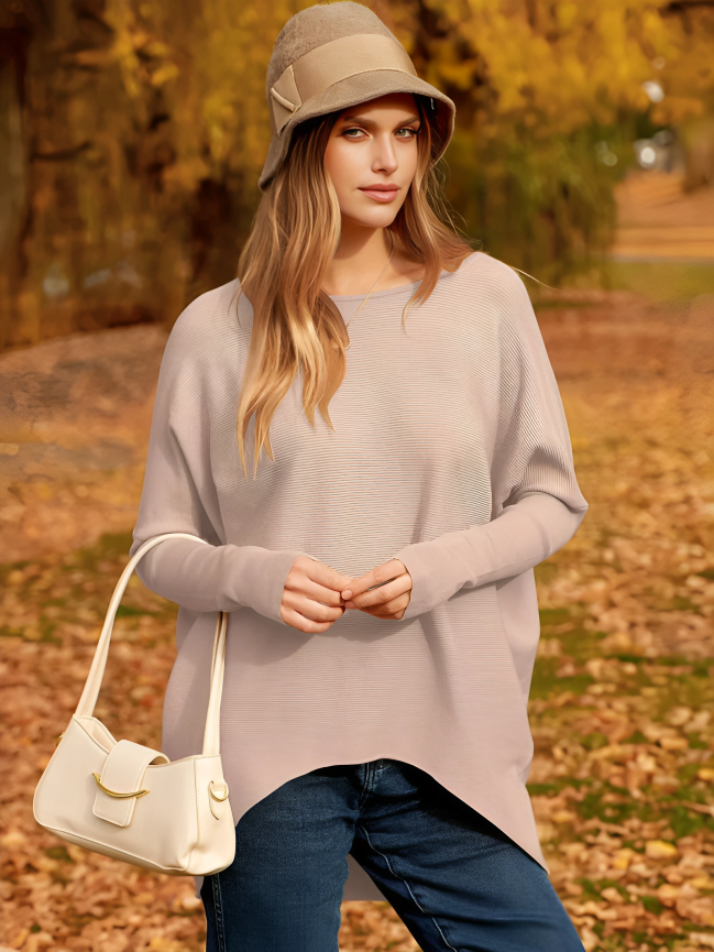 Femme en pull beige et chapeau, posant dans un parc automnal avec feuilles jaunes, portant un sac à main blanc. Mode automne, style décontracté.
