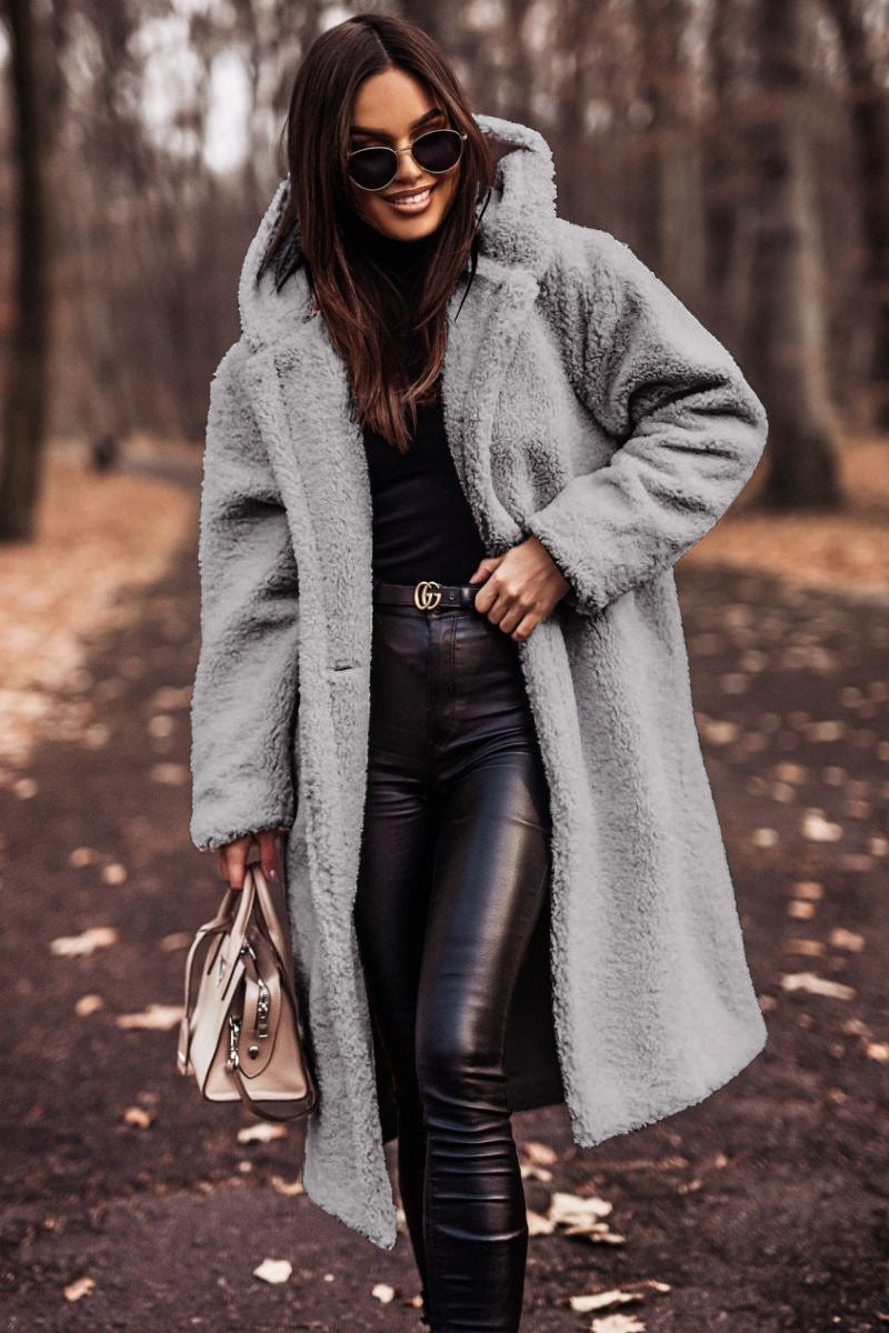 Femme portant un manteau en fausse fourrure gris, lunettes de soleil, pantalon en cuir noir, tenant un sac à main beige, marchant dans un parc automnal.