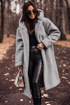 Femme portant un manteau en fausse fourrure gris, lunettes de soleil, pantalon en cuir noir, tenant un sac à main beige, marchant dans un parc automnal.