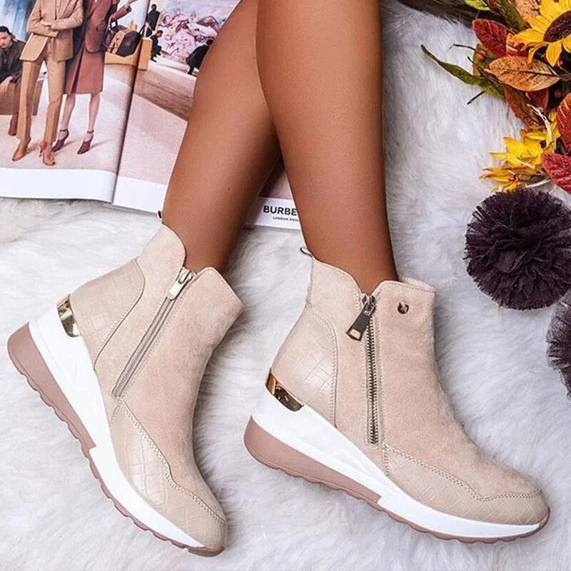 Bottes beiges en daim avec fermeture éclair, semelle compensée blanche et détails dorés, posées sur un tapis blanc, à côté d'un magazine ouvert.