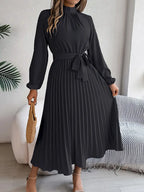 Robe longue noire plissée élégante avec manches longues, ceinture nouée à la taille, portée par une femme tenant un sac en osier.