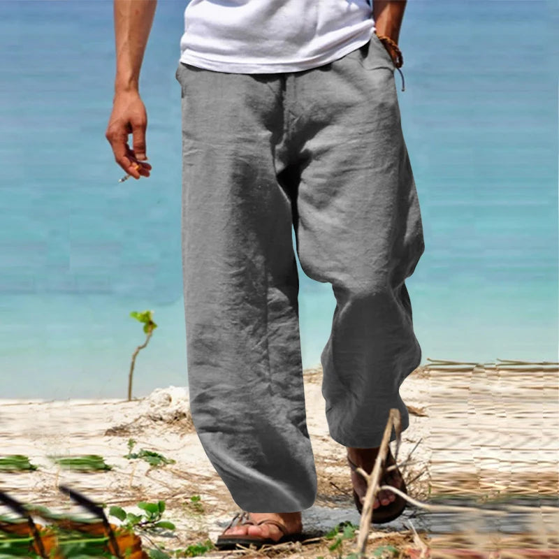 Homme portant un pantalon en lin gris, marchant sur une plage avec vue sur la mer, vêtu de sandales et d'un t-shirt blanc. Mode estivale décontractée.