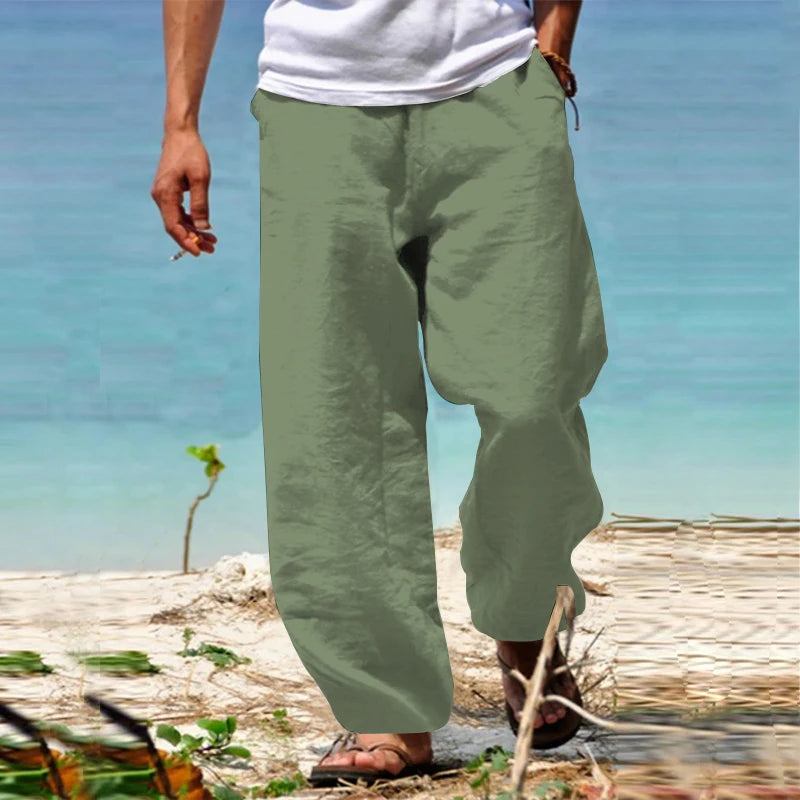 Homme portant un pantalon en lin vert kaki sur une plage, pieds nus, avec une mer turquoise en arrière-plan. Mode estivale décontractée.