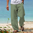 Homme portant un pantalon en lin vert kaki sur une plage, pieds nus, avec une mer turquoise en arrière-plan. Mode estivale décontractée.