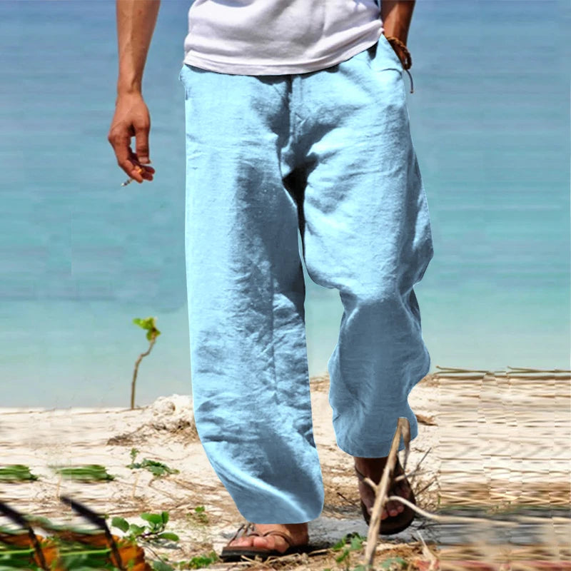 Homme portant un pantalon en lin bleu clair sur une plage, pieds nus dans le sable, mer en arrière-plan. Mode estivale décontractée, vacances plage.