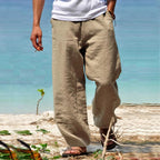 Homme portant un pantalon en lin beige, marchant sur une plage avec vue sur la mer, style décontracté estival, mode masculine plage.