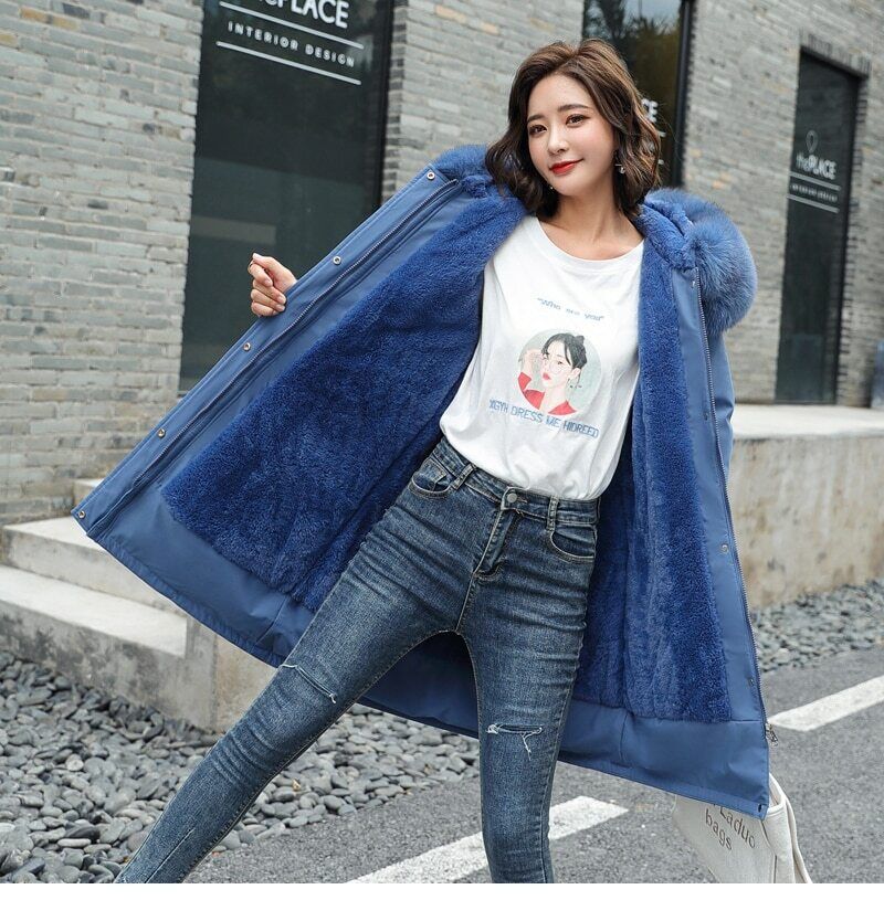 Femme portant un manteau bleu avec capuche en fourrure, t-shirt blanc et jean, posant devant un mur de briques grises. Mode hiver tendance.