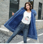 Femme portant un manteau bleu avec capuche en fourrure, t-shirt blanc et jean, posant devant un mur de briques grises. Mode hiver tendance.