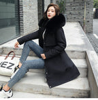 Femme en manteau noir avec col en fourrure, assise sur des marches en béton, portant un jean slim et des baskets blanches, sac blanc à côté.