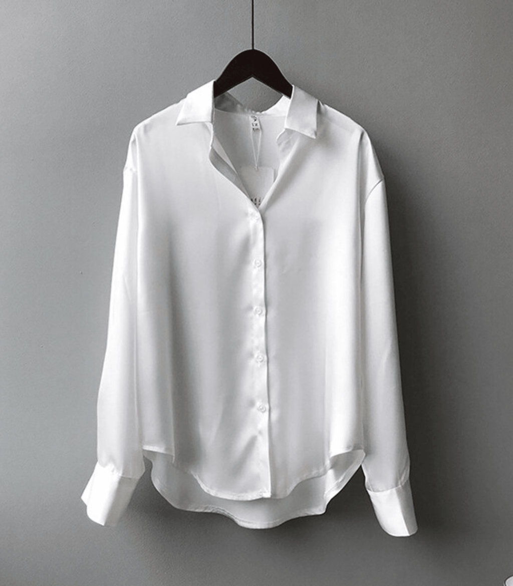 Chemise blanche en satin pour femme, manches longues, col classique, suspendue sur un cintre noir, fond gris. Mode élégante et minimaliste.
