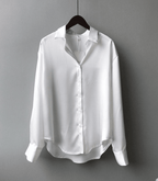 Chemise blanche en satin pour femme, manches longues, col classique, suspendue sur un cintre noir, fond gris. Mode élégante et minimaliste.