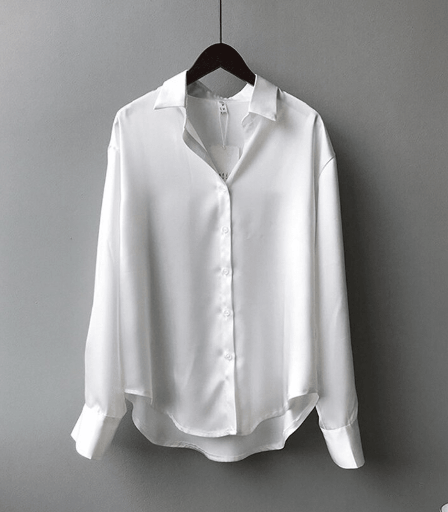 Chemise blanche en satin pour femme, manches longues, col classique, suspendue sur un cintre noir, fond gris. Mode élégante et minimaliste.