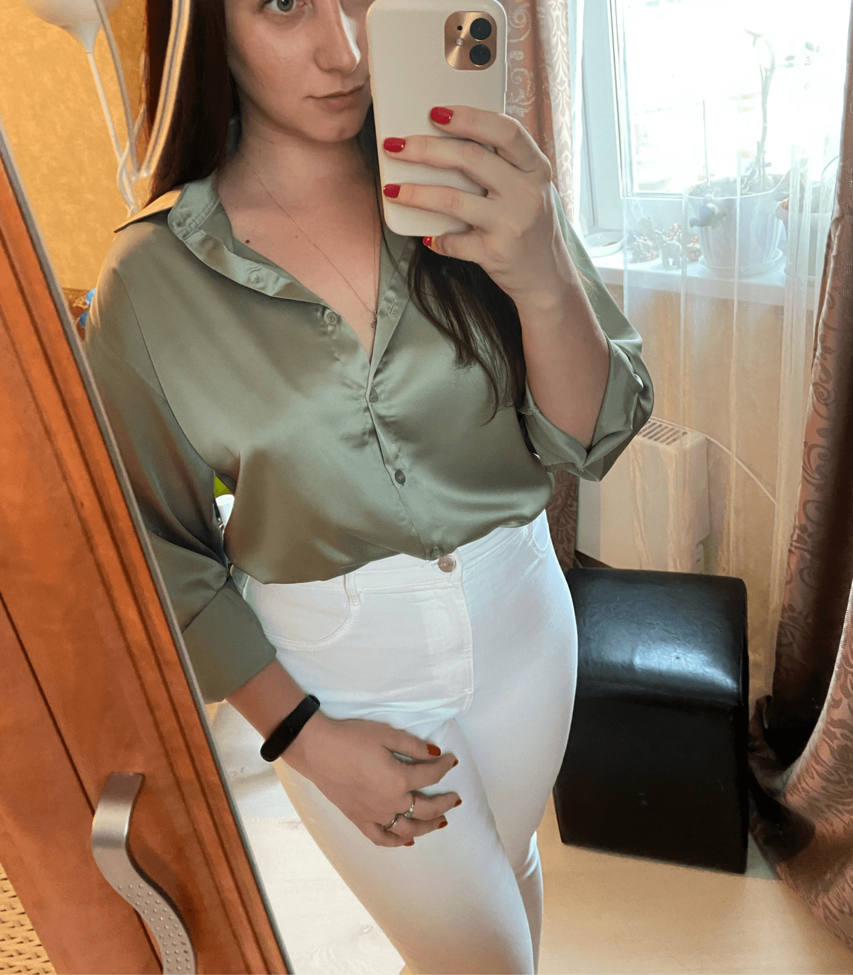 Femme prenant un selfie dans un miroir, portant une chemise verte et un pantalon blanc, tenant un smartphone doré. Mode décontractée, intérieur lumineux.