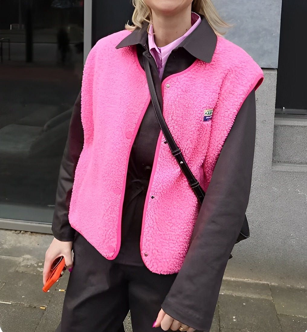 Femme portant un gilet rose vif en laine polaire sur une chemise noire, tenant un smartphone orange. Mode urbaine, tendance automne-hiver.