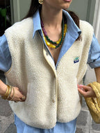 Femme portant un gilet en laine bouclée blanc sur une chemise bleue, colliers colorés, sac tressé jaune, bracelets dorés. Mode automne chic.