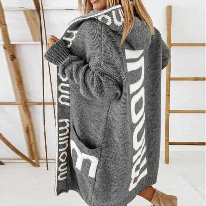 Femme portant un long cardigan gris à capuche avec motifs blancs, style décontracté, tricoté, idéal pour l'automne et l'hiver.
