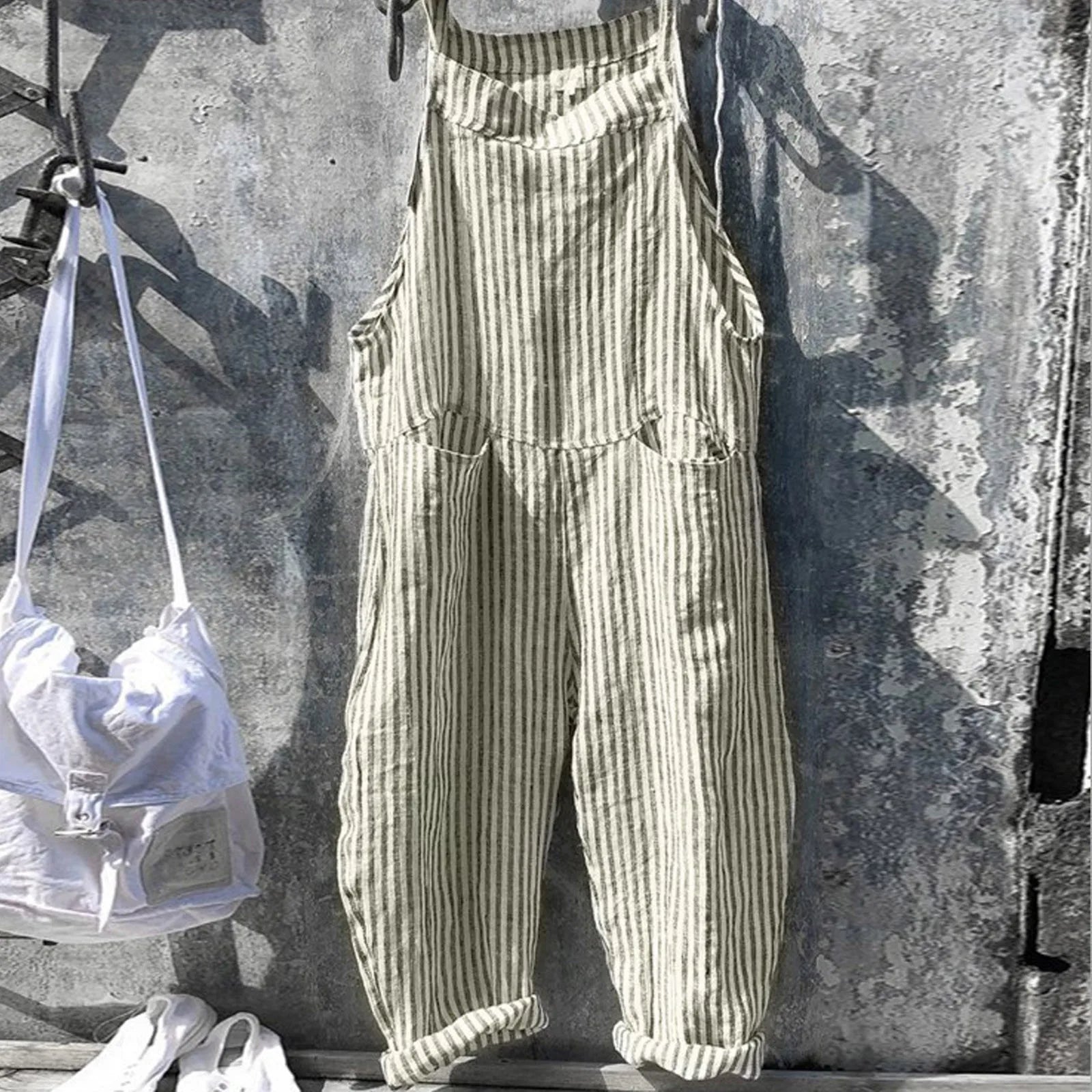 Salopette rayée beige suspendue sur un mur en béton, accompagnée d'un sac à dos blanc et de baskets blanches. Mode décontractée et tendance.