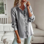 Femme portant une chemise rayée grise à volants, manches longues, posant dans un salon moderne. Mode féminine élégante, tenue décontractée.