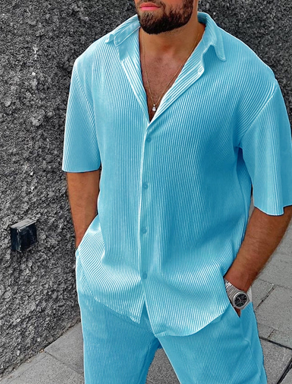 Homme portant une chemise et un pantalon bleu clair plissés, style décontracté, avec une montre argentée, sur fond de mur texturé gris.