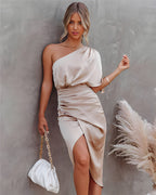 Femme en robe beige asymétrique élégante, tenant un sac blanc à chaîne dorée, posant contre un mur gris avec des herbes pampas en arrière-plan.
