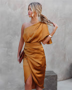 Femme en robe asymétrique satinée dorée, tenant une pochette marron, posant contre un mur texturé. Mode élégante, tenue de soirée chic.