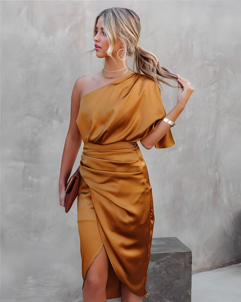 Femme en robe asymétrique satinée dorée, tenant une pochette marron, posant contre un mur texturé. Mode élégante, tenue de soirée chic.