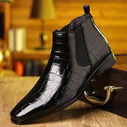 Bottine Chelsea en cuir noir verni à motif croco, posée sur une surface en bois, avec fond flou de bibliothèque. Chaussure élégante pour homme.