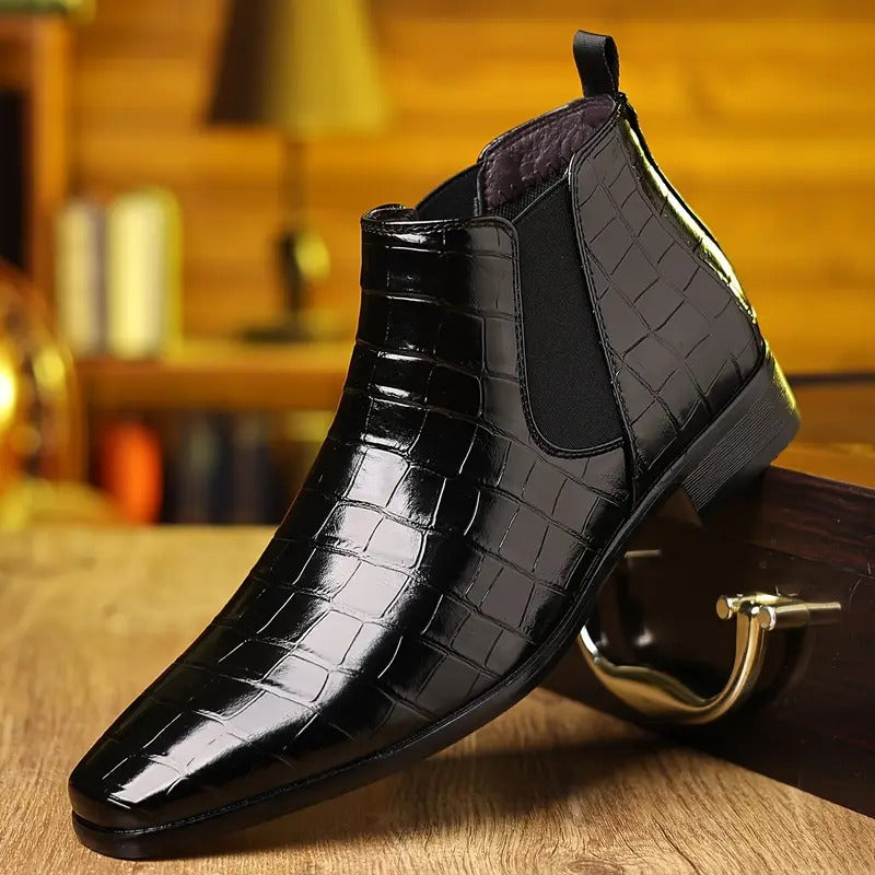 Bottine Chelsea en cuir noir verni à motif croco, posée sur une surface en bois, avec fond flou de bibliothèque. Chaussure élégante pour homme.
