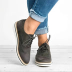 Chaussures derby en cuir noir pour femme, style décontracté, portées avec un jean bleu retroussé, sur fond de plancher en bois blanc.