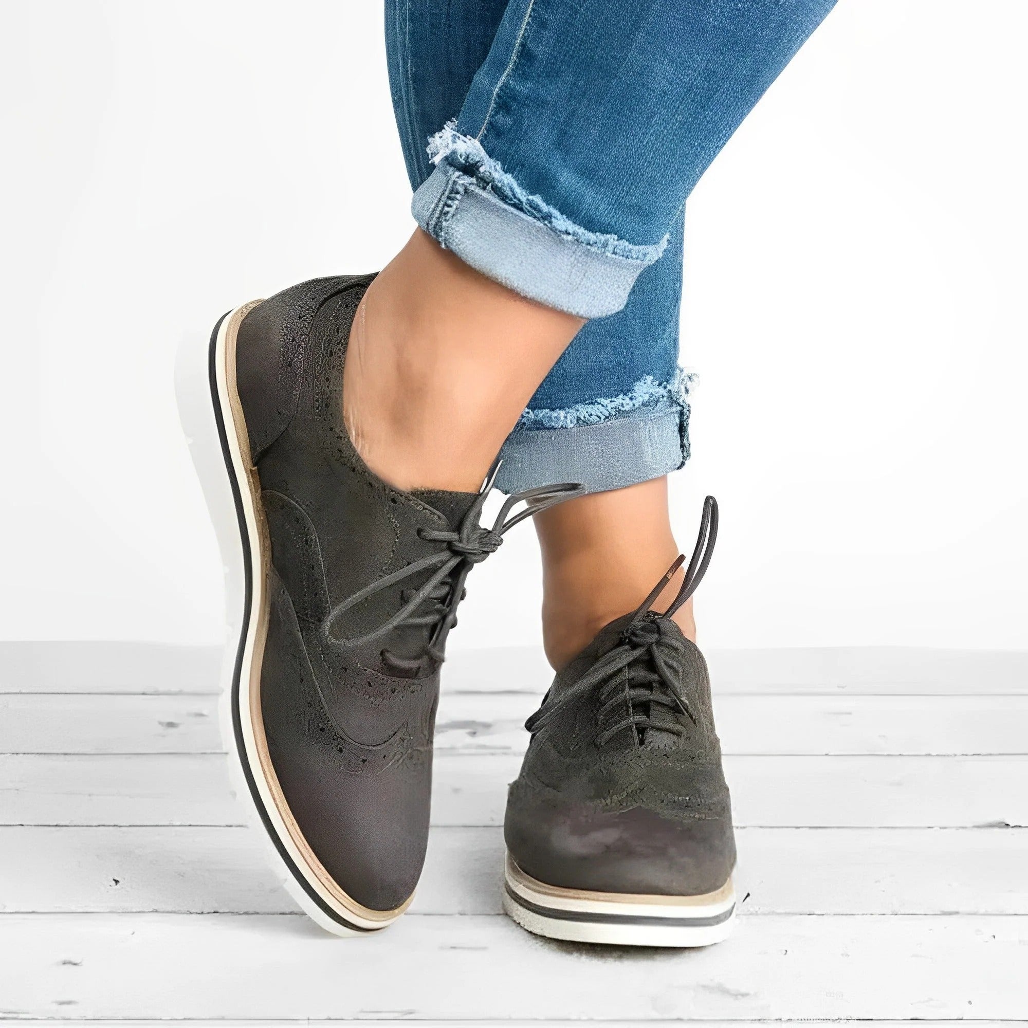 Chaussures derby en cuir noir pour femme, style décontracté, portées avec un jean bleu retroussé, sur fond de plancher en bois blanc.